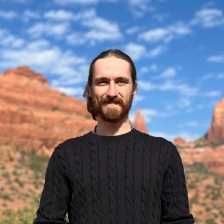 Man with a brown beard and brown hair pulled back into a bun. Wearing a black long sleeved shirt. Top half visible. The background is sandstone cliffs in Sedona, Arizona with a blue sky and scattered cloud.